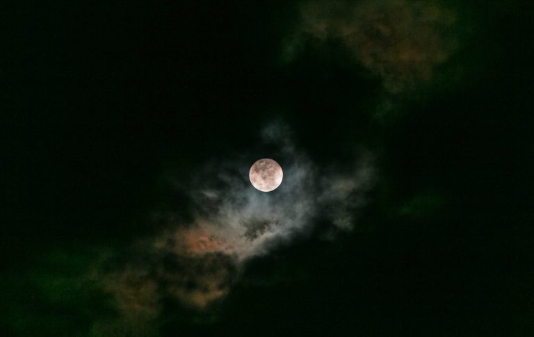 Full Moon in a dark sky viewed through the clouds. The clouds appear reddish next to the moon